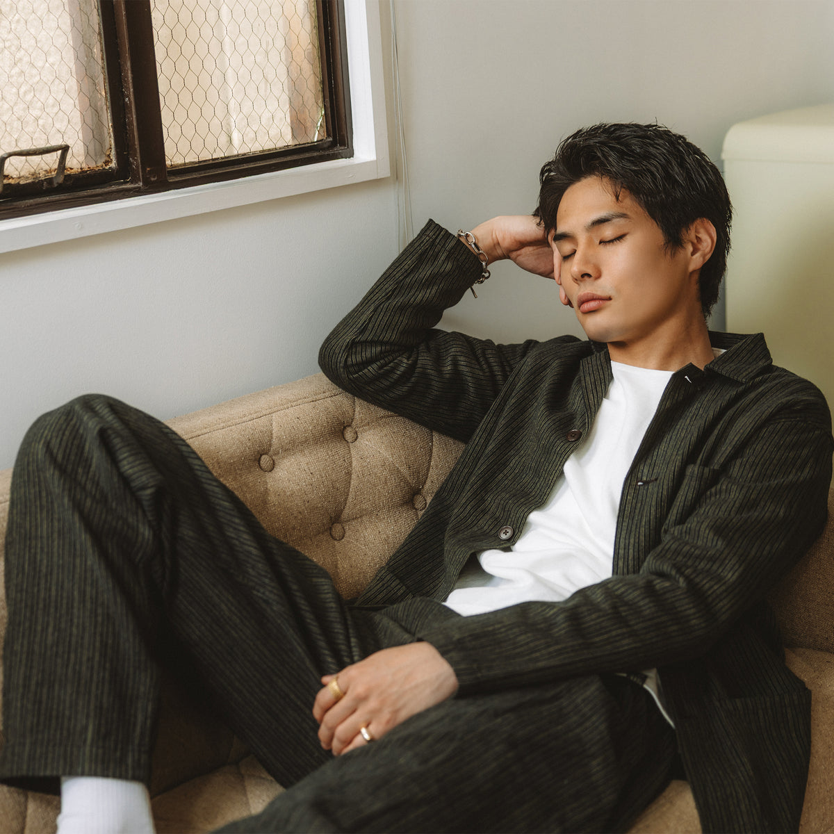 PREQUEL P1008Bのストライプパンツと同柄のジャケットを白いTシャツに合わせ、ソファに腰掛けたモデルの着用写真。