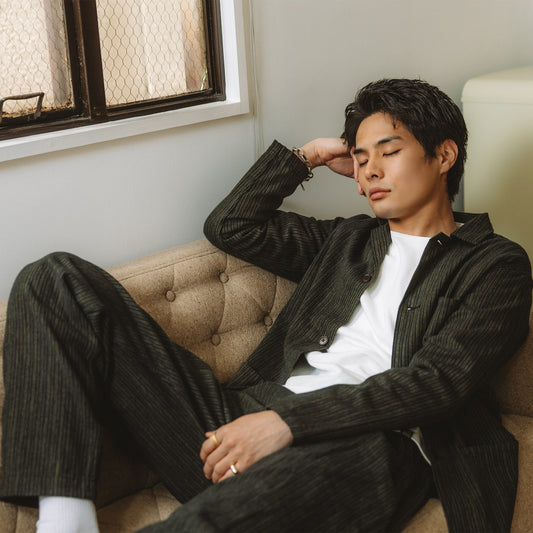 PREQUEL P1008Bのストライプパンツと同柄のジャケットを白いTシャツに合わせ、ソファに腰掛けたモデルの着用写真。